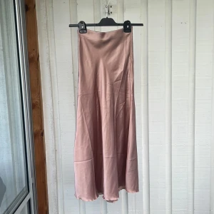 Ljusrosa satinkjol i lång modell - Superfin ljusrosa långkjol i satin med mjukt fall och lätt glansig yta. Kjolen har en enkel och stilren design utan synliga detaljer, perfekt för att skapa en chic look.