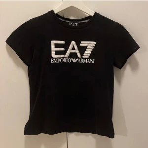 Svart EA7 Emporio Armani t-shirt - Svart t-shirt från EA7 Emporio Armani med stor vit logotyp på bröstet. Klassisk rund hals och korta ärmar. Snygg och enkel design som passar perfekt till jeans eller shorts. Tillverkad i mjuk bomull för skön komfort.