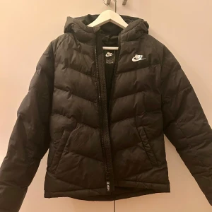 Svart pufferjacka från Nike - Svart pufferjacka från Nike med huva och vit logga på bröstet. Jackan har quiltad design och dragkedja framtill.