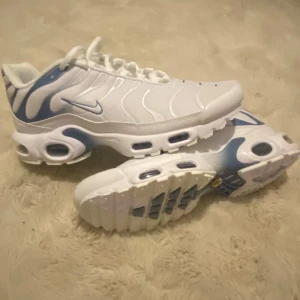 Nike Air Max Plus vita/blå sneakers - Nike Air Max Plus sneakers i vitt med blå detaljer och vågigt mönster på sidan. Snygg design med synliga luftkuddar i sulan, snörning och rund tå. Ovandelen är i syntet och mesh för bra komfort och ventilation. Perfekt för dig som gillar sportig och modern stil.