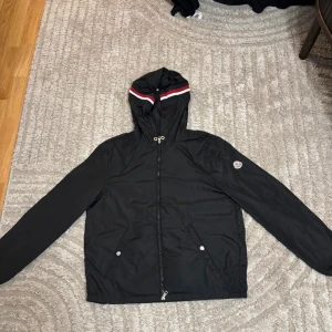 Svart vindjacka från Moncler - Svart vindjacka från Moncler med huva som har röd och vit rand. Jackan har dragkedja framtill, två fickor med tryckknappar och Moncler-logga på ärmen. Perfekt för dig som vill ha en stilren och sportig look.