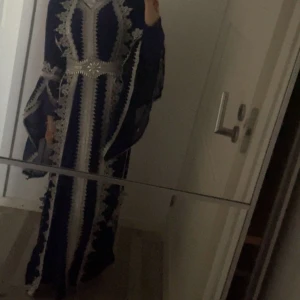 Blå långklänning med silverbroderi - Säljer en blå långklänning med vackra silverbroderier och markerad midja med ett brett bälte. Klänningen har vida ärmar och detaljerade mönster längs hela plagget, vilket ger en lyxig och elegant känsla. Perfekt för dig som vill sticka ut med en unik look. Den är custom made.  Super fint kurdisk/turisk klänning 