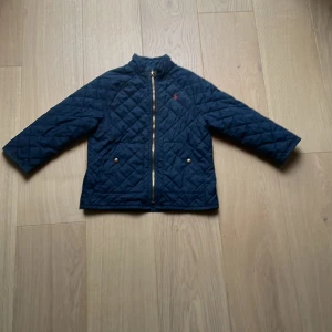 Mörkblå Ralph lauren jacka för småbarn. - Quiltad mörkblå jacka från Polo Ralph Lauren med guldfärgad dragkedja och två fickor med tryckknappar. Jackan har lång ärm och stårkrage, tillverkad i polyester. Perfekt för kyliga dagar och snygg till jeans eller chinos.