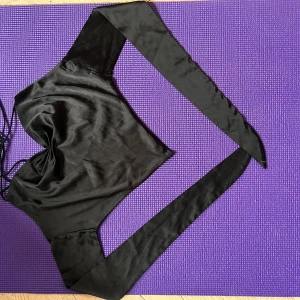 Svart omlottopp i satin från Cider - Supersnygg svart omlottopp från Cider i glansigt satinliknande material. Toppen har smala axelband, offshoulder-långa ärmar och v-ringning med lätt rynk framtill. Sitter croppad och knyts i ryggen för en trendig look.