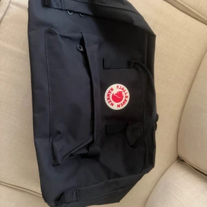 Svart Fjällräven Kånken ryggsäck - Klassisk svart Fjällräven Kånken ryggsäck med ikonisk rund röd och vit logga framtill. Väskan har ett stort huvudfack, en mindre ficka med dragkedja på framsidan och justerbara axelremmar. Tillverkad i slitstark polyester med metallknappar och detaljer.