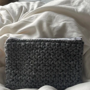 Grå virkad necessär med dragkedja - En handgjord grå necessär med virkad utsida i blommigt mönster och vit dragkedja. Insidan är fodrad med grått tyg med små prickar i olika färger. Perfekt för att bära småsaker eller smink.