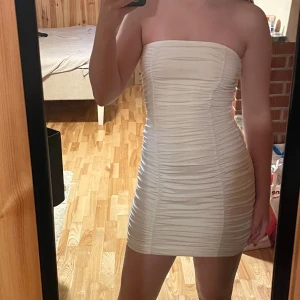 Vit axelbandslös kortklänning - Säljer en vit, axelbandslös kortklänning med snygg rynkad design. Klänningen är tight och har en clean, modern look som verkligen framhäver figuren. Perfekt för dig som gillar minimalistisk stil med en twist. Materialet känns mjukt och stretchigt mot huden.