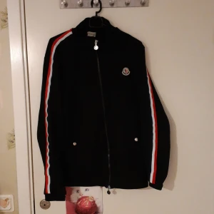 Svart Moncler vindjacka med ränder - Svart vindjacka från Moncler med dragkedja och coola röda och vita ränder längs ärmarna. Jackan har Moncler-logga på bröstet och två fickor framtill med knappar. Perfekt för dig som vill ha en sportig och stilren look.