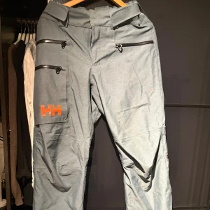 Grå skidbyxor från Helly Hansen - Snygga grå skidbyxor från Helly Hansen med orange HH-logga på benet. Utrustade med flera dragkedjefickor, förstärkta benslut och RECCO-reflektor för extra säkerhet. Perfekta för vinterns äventyr och outdoor-aktiviteter.