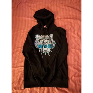 Kenzo - Säljer en svart hoodie från Kenzo med ikoniskt broderat tigertryck i blått, vitt och grått på bröstet. Tröjan har huva med snörning och en klassisk känguruficka framtill. Perfekt för dig som gillar streetwear och vill sticka ut med en cool design.