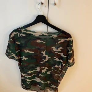 Camo mesh topp från Midnight Fashion - Säljer en transparent mesh topp från Midnight Fashion med camouflage-mönster i grönt, brunt och beige. T-shirten har korta ärmar och en loose fit, perfekt för att skapa en edgy look. Superlätt och luftig, snygg att styla lager på lager.