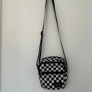 Svartvit rutig axelväska från Stuhf - Axelväska med svartvitt rutigt mönster och justerbar rem. Väskan har två fack med dragkedja och en patch med text framtill. Tillverkad i bomullstyg, perfekt för att bära dina viktigaste grejer. Snygg och unik design som sticker ut.