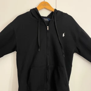 Svart hoodie från Polo Ralph Lauren - Svart hoodie från Polo Ralph Lauren med klassisk broderad logga i vitt på bröstet. Tröjan har huva med snörning, dragkedja framtill och två rymliga fickor. Tillverkad i mjuk bomull och har långa ärmar för extra komfort. Pris kan diskuteras privat 