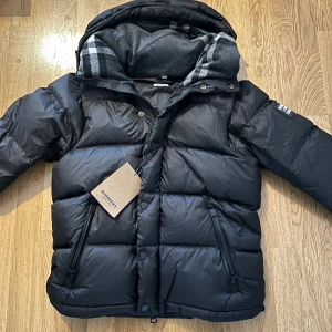 Burberry Jacka - Svart pufferjacka från Burberry med avtagbara ärmar, vilket gör att den kan användas som väst. Jackan har huva med klassiskt rutigt Burberry-foder, dragkedjor och tryckknappar framtill samt två sidofickor med dragkedja. Stor Burberry-patch på ena ärmen.