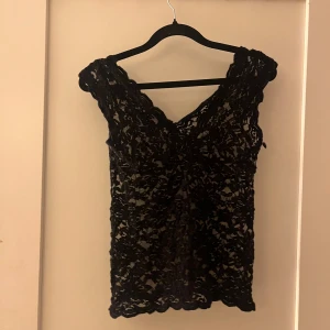 Svart spetsblus med v-ringning - Säljer en svart blus i spets med v-ringad hals och ärmlös design. Blusen har ett genombrutet mönster och romantisk känsla, perfekt för dig som gillar detaljer och vill sticka ut. Snygg att styla med jeans eller kjol för en cool look.