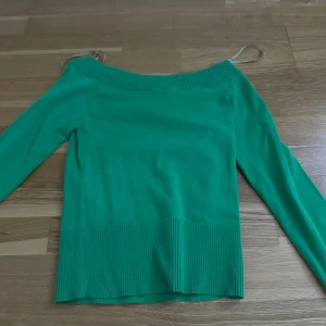 Grön offshoulder stickad tröja - Snygg grön stickad tröja i offshoulder-modell med långa ärmar. Tröjan har bred ribbad kant vid halsringning och nederkant som ger en cool och modern vibe. Perfekt för dig som gillar färg och vill sticka ut lite extra.
