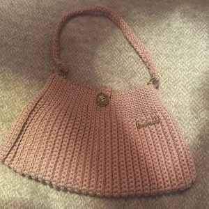 Handgjord rosa stickad handväska - Supersöt handväska i stickad design med mjuk rosa färg. Väskan har ett guldfärgat spänne och en matchande axelrem. Perfekt accessoar för dig som vill ha något unikt och personligt. Liten metallskylt med texten 'Handmade' på framsidan.