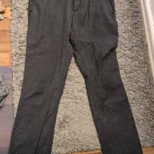 Grå rutiga chinos från Emilio Pants stl 52 - Snygga grå chinos från Emilio Pants med diskret rutigt mönster. Byxorna har raka ben, bälteshällor och klassisk passform. Tillverkade i en skön mix av bomull, polyester och elastan för extra komfort. Perfekta för en stilren look.