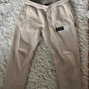 Beige mjukisbyxor från Hoodrich - Beiga mjukisbyxor från Hoodrich med broderad logga på låret. Byxorna har snörning i midjan och två fickor framtill. Mjukt och skönt material som passar perfekt för chill dagar. Snygg streetwear-vibe och relaxed fit.