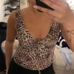 Leopardmönstrad linnetopp med snörning - Säljer en snygg leopardmönstrad linnetopp med djup v-ringning och snörning framtill. Toppen är ärmlös och har en figurnära passform som framhäver siluetten. Perfekt för dig som gillar att sticka ut med coola mönster och detaljer.