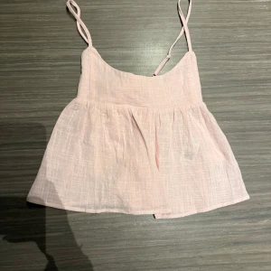 Ljusrosa linnetopp med rosett från Nelly - Supersöt ljusrosa linnetopp från Nelly med tunna axelband och en stor rosett i ryggen. Toppen är lätt och luftig med en svagt strukturerad yta och har en feminin, något utsvängd form. Perfekt för varma dagar och sommarhäng.