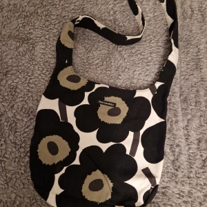 Marimekko axelväska med blommönster - Snygg axelväska från Marimekko med ikoniskt svart och beige blommönster. Väskan har en bred axelrem, magnetknäppning och en praktisk innerficka med dragkedja. Perfekt storlek för vardagsgrejer och ger en cool touch till din outfit.