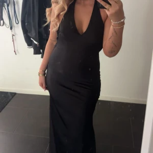 Svart långklänning med öppen rygg - Supersnygg svart långklänning i satinliknande material med djup v-ringning och öppen rygg. Klänningen har halterneck med breda band som knyts i nacken och en tight passform som framhäver figuren. 