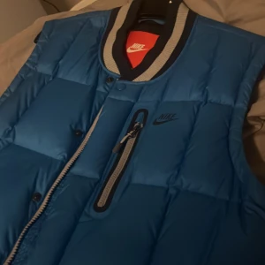 Blå dunväst från Nike - Helt ny oanvänd snygg blå dunväst från Nike med svart logga på bröstet. Västen har quiltad design, dragkedja och knappar framtill samt en randig ribbad krage i svart och grått. Praktisk bröstficka med dragkedja och sportig look. Perfekt för lager-på-lager under kyliga dagar. Priset går att diskutera 🤝