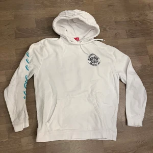 Vit Santa Cruz hoodie med tryck - Vit hoodie från Santa Cruz med huva och känguruficka. Tryck med Santa Cruz-logga på bröstet och stort färgglatt tryck med japanska tecken på ryggen. Blått tryck längs ena ärmen. Perfekt för dig som gillar streetwear och skate. Tröjan är väldigt oanvänd och då i bra skick👍