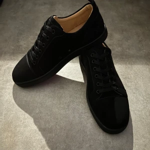 Christian Louboutin svarta sneakers mocka - Snygga svarta sneakers från Christian Louboutin i mocka med klassisk röd sula och broderad logga baktill. Skorna har rund tå, svarta skosnören och lyxig innersula i skinn. Perfekta för dig som vill sticka ut med exklusiv streetstyle.