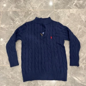 Marinblå stickad tröja Polo Ralph Lauren - Säljer en marinblå stickad tröja från Polo Ralph Lauren med klassisk kabelstickning och röd broderad logga på bröstet. Tröjan har half zip och ribbade muddar vid hals, ärmslut och nederkant. Perfekt för dig som gillar stilren och tidlös design.