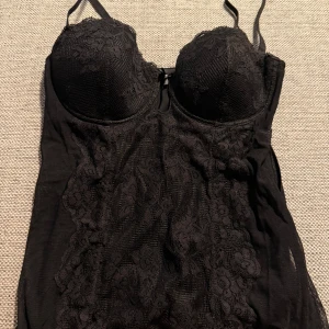 Svart spetskorsett med kupor - Säljer en svart korsett med vadderade kupor och tunna axelband från intimissimi. Framsidan är täckt av snygg spets med blommönster och meshdetaljer. 