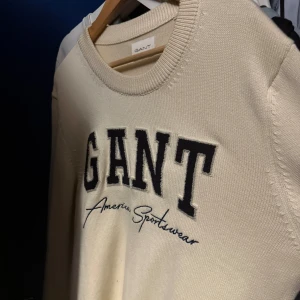 Vit stickad tröja från GANT - Snygg vit stickad tröja från GANT med stor broderad logga i svart och blått på bröstet. Rund hals och klassisk passform, perfekt för dig som gillar preppy stil. Tröjan har ribbade muddar och känns riktigt mjuk och skön.