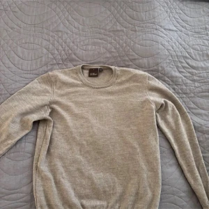 Beige ulltröja från Oscar Jacobson - Klassisk beige tröja från Oscar Jacobson i mjuk ull. Tröjan har rund halsringning och långa ärmar, perfekt för lager-på-lager. Tillverkad i Italien och har en stilren, tidlös design som passar till mycket.