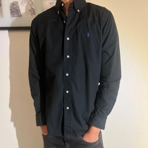 Svart skjorta från Ralph Lauren - Klassisk svart skjorta från Ralph Lauren med custom fit-passform. Skjortan har vita knappar, button-down krage och lila broderad logga på bröstet. Tillverkad i bomull och har långa ärmar. Perfekt för en stilren look.