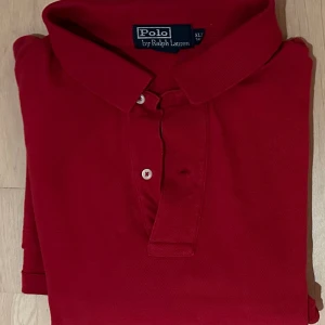 Röd pikétröja från Polo Ralph Lauren - Riktigt snygg röd pikétröja från Polo Ralph Lauren med klassisk krage och två vita knappar framtill. Tröjan har korta ärmar och den ikoniska broderade loggan på bröstet. Tillverkad i mjuk bomull som är skön mot huden.