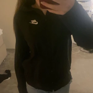 Svart Nike hoodie med dragkedja - Svart hoodie från Nike med vit logga på bröstet. Tröjan har lång ärm, huva och dragkedja framtill. Perfekt för chill dagar eller när du vill ha en sportig look. Materialet känns mjukt och bekvämt mot huden.