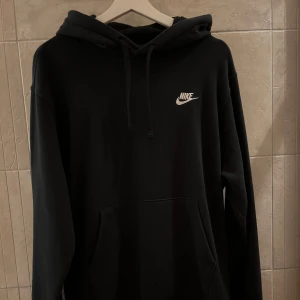 Svart Nike hoodie med huva - Svart hoodie från Nike med klassisk logga i vitt på bröstet. Tröjan har huva med snörning och stor magficka framtill. Mjuk insida och långärmad modell, perfekt för chill eller träning. Snygg och enkel stil som funkar året runt.