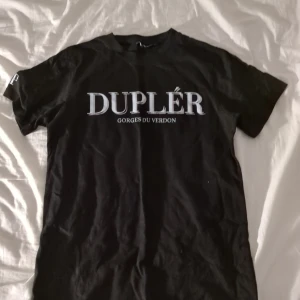 Svart t-shirt från Duplér - Svart t-shirt från Duplér med vit text 'DUPLÉR GORGES DU VERDON' tryckt på bröstet. Klassisk passform med rund hals och korta ärmar. Tillverkad i mjuk bomull, perfekt för en clean och stilren look.