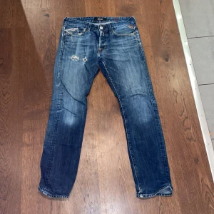 Replay Jeans - Snygga blå jeans från Replay med slitningar och lagom tvättad look. Klassisk femficksmodell med raka ben och normal passform. Tydliga Replay-detaljer vid fickan och robust denimkvalitet. Perfekta för dig som gillar en avslappnad stil. Hål i skrevet därav priset, det syns inte vid användning och kan enkelt repareras.