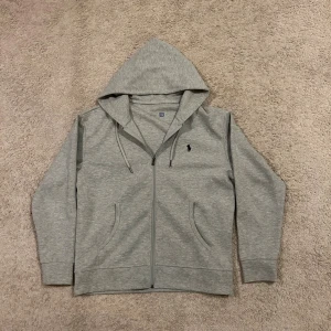 Grå hoodie från Polo Ralph Lauren - Snygg grå hoodie från Polo Ralph Lauren med dragkedja och huva. Tröjan har snörning vid huvan, två fickor fram och den klassiska Polo-loggan broderad på bröstet. Perfekt för chill dagar och enkel att matcha med jeans eller joggers.
