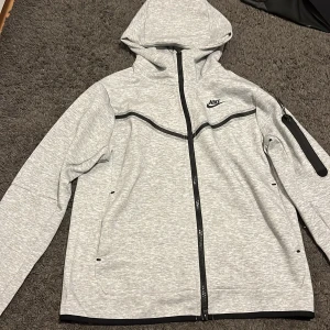 Grå vindjacka från Nike med huva - Snygg grå vindjacka från Nike med svart logga på bröstet. Jackan har hel dragkedja, huva och svarta detaljer längs sömmarna. Praktisk ficka med dragkedja på ärmen och två sidofickor. Perfekt för dig som gillar sportig stil.