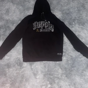Svart hoodie Supply & Demand NYC - Svart hoodie från Supply & Demand New York City i storlek M. Tryck på bröstet med texten 'Supply & Demand' i vit och guld. Klassisk huva med snörning och känguruficka framtill. Perfekt för dig som gillar streetwear och snygga detaljer. 