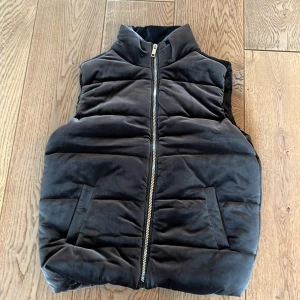 Grafitgrå quiltad dunväst med dragkedja - Grafitgrå dunväst i storlek M med quiltad design och hög krage. Västen har två snedställda fickor framtill och stängs med en silverfärgad dragkedja. Materialet är mjukt och har en matt finish, vilket ger en stilren look. Perfekt att slänga över en hoodie för extra värme. Västen är helt ny och säljs till halva priset på grund av fel storlek. 