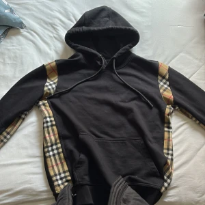 Svart Burberry hoodie med rutiga detaljer - Svart hoodie från Burberry med klassiska rutiga paneler i beige, svart och rött längs sidorna och ärmarna. Tröjan har huva med snörning, känguruficka och ribbade muddar. Perfekt för dig som vill ha en lyxig streetwear-look med ikonisk Burberry-vibe.