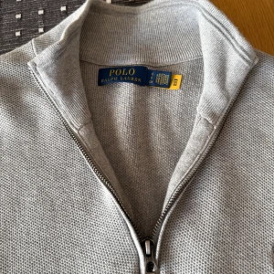 Grå half zip tröja från Polo Ralph Lauren - Snygg grå stickad tröja från Polo Ralph Lauren med half zip och hög krage. Tröjan har långärmad passform och diskret logga broderad på bröstet. Perfekt för dig som gillar stilrena och klassiska plagg med en modern touch. Tröjan är i toppskick, använd endast ett fåtal gånger