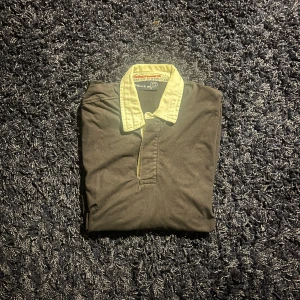 Svart henley från Pelle P  - Svart henley från Pelle P med vit krage och broderade detaljer på ärmen, inklusive koordinater och logotyp. Skjortan har korta ärmar och knappar vid halsen. Materialet är mjuk bomull, perfekt för en chill och stilren look.