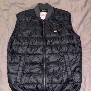 Svart dunväst från Tommy Jeans - Svart quiltad dunväst från Tommy Jeans med dragkedja framtill och ribbad krage. Västen har två fickor på sidorna och en liten logga på bröstet. Perfekt för lager-på-lager och enkel att matcha med olika outfits. Är använd en höst och är i bra skick.