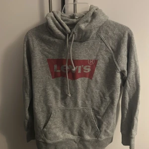 Grå hoodie från Levi's med logga - Säljer en grå hoodie från Levi's med klassisk röd logga framtill. Tröjan har huva med snörning och en stor magficka. Perfekt för dig som gillar en avslappnad och sportig stil.