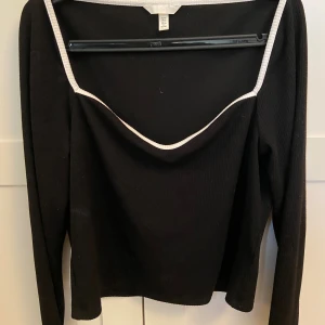 Svart långärmad topp från H&M XL - Svart långärmad topp från H&M i storlek XL med vit kant runt den breda, djupa halsringningen. Toppen har en ribbad struktur och är croppad i modellen, vilket ger en modern och stilren look. Perfekt för dig som gillar kontraster och enkel design.